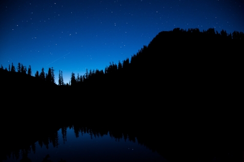 A night-scape of a lake surrounded by a forst and mountains at a distance